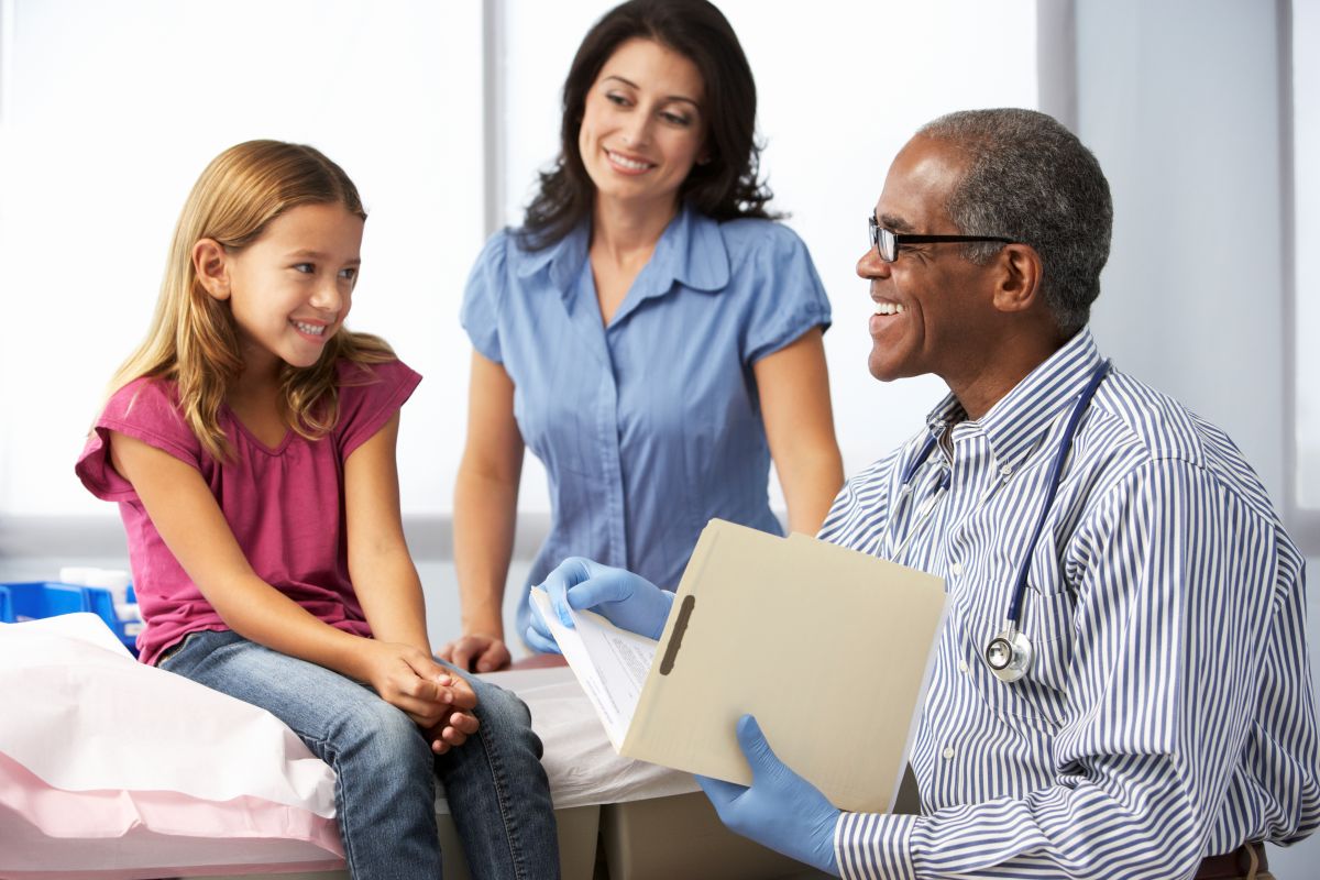 African American male doctor treating a young white girl with her mother in a clinical setting.